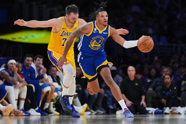 A Golden State Warriors player dribbles the basketball past a Los Angeles Lakers defender during a game, with spectators visible in the background.
